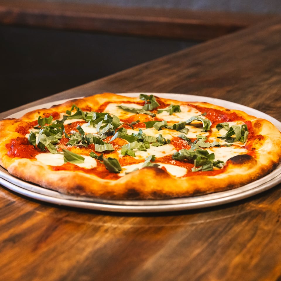 A freshly baked Margherita pizza with basil and mozzarella on a metal pan, on a wooden table.